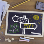 A chalkboard shows two directional signs labeled "Should I stay?" and "Should I go?"—a nod to couples considering divorce or seeking discernment counseling—with drawings of a sun and tree, surrounded by office supplies and coins on a wooden desk.