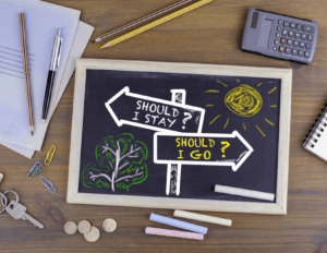 A chalkboard shows two directional signs labeled "Should I stay?" and "Should I go?"—a nod to couples considering divorce or seeking discernment counseling—with drawings of a sun and tree, surrounded by office supplies and coins on a wooden desk.