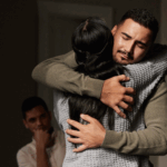 Two people embrace tightly, eyes closed, while a third person sits in the background watching them, reflecting on healing and coping during their journey through the 3 C's of Grief.