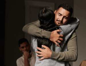 Two people embrace tightly, eyes closed, while a third person sits in the background watching them, reflecting on healing and coping during their journey through the 3 C's of Grief.
