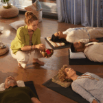 At a wellness retreat at Bay Area CBT Center, a woman kneels on the floor with a singing bowl while four people lie on mats, eyes closed, participating in a calming sound meditation session.