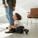 A young boy sits on the floor, hugging an adult’s leg and looking up at them, reflecting attachment styles. The adult stands in a bright room with a wooden bench in the background.