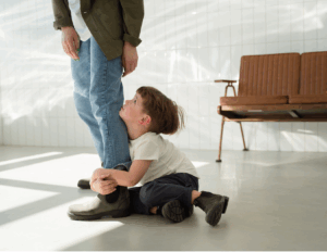 A young boy sits on the floor, hugging an adult’s leg and looking up at them, reflecting attachment styles. The adult stands in a bright room with a wooden bench in the background.