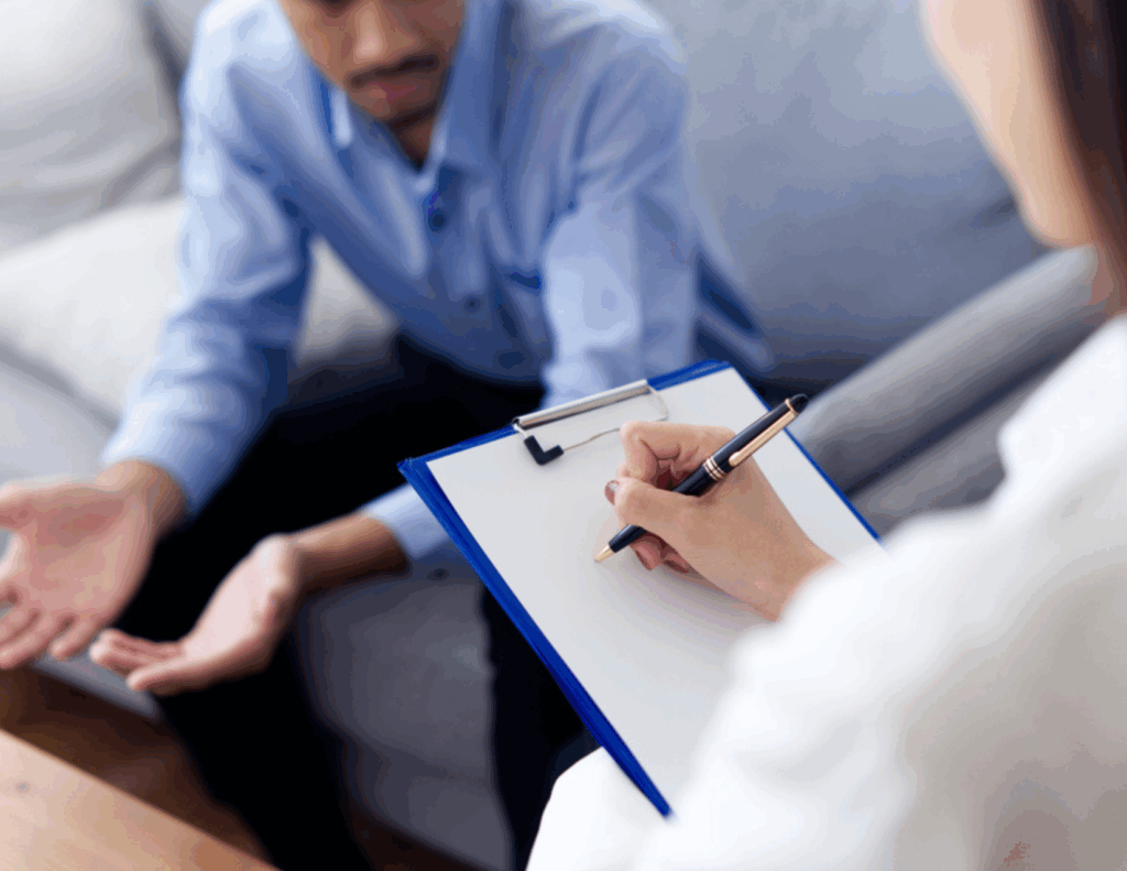 A person holding a clipboard takes notes during an Intensive Outpatient Care session, while another individual sits on a couch engage in conversation about an IOP program near me.