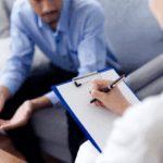 A person holding a clipboard takes notes during an Intensive Outpatient Care session, while another individual sits on a couch engage in conversation about an IOP program near me.