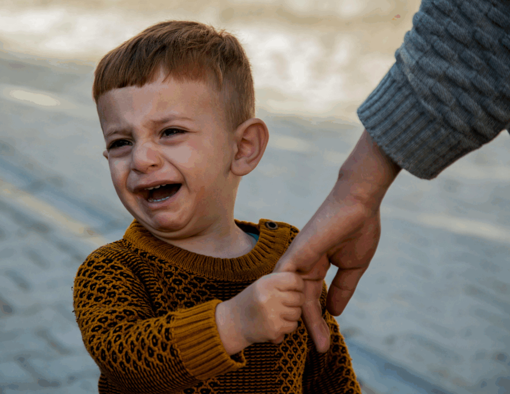 A young child wearing a brown sweater is crying while holding an adult's hand outdoors, possibly displaying signs of a disorganized attachment style and highlighting the need to explore causes and solutions.