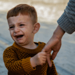 A young child wearing a brown sweater is crying while holding an adult's hand outdoors, possibly displaying signs of a disorganized attachment style and highlighting the need to explore causes and solutions.
