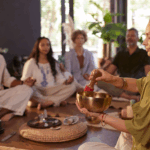 A group of people sit cross-legged in a circle, eyes closed, while one person strikes a singing bowl in a calm, indoor setting—an atmosphere ideal for rewiring patterns at mental health wellness retreats.