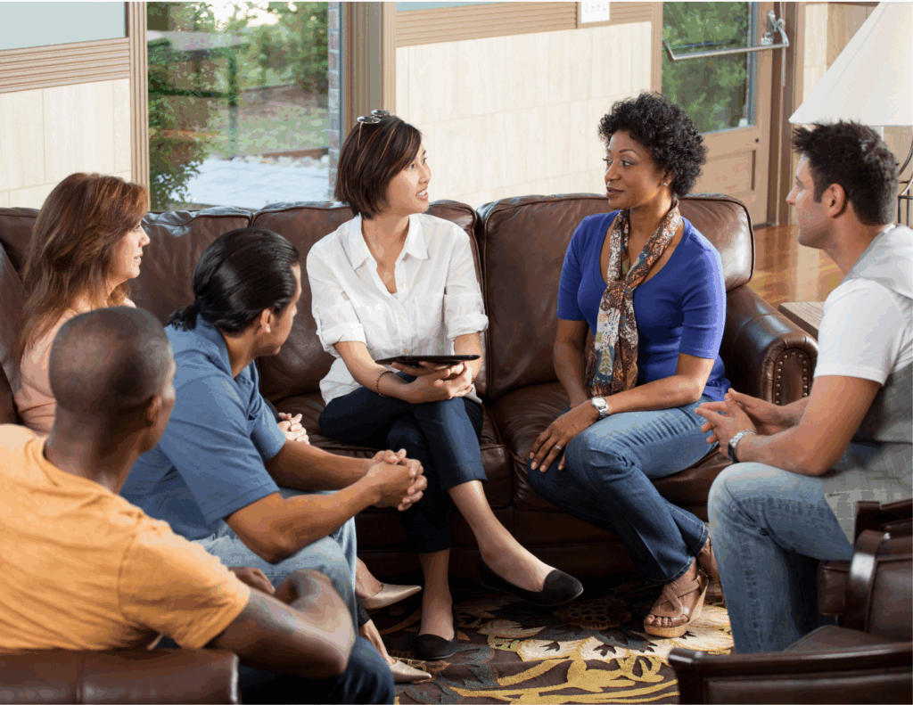 Six adults sit in a circle on couches and chairs, engaged in a group therapy session in a living room setting. One woman in the center holds a tablet and appears to be leading an effective healing discussion.