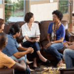 Six adults sit in a circle on couches and chairs, engaged in a group therapy session in a living room setting. One woman in the center holds a tablet and appears to be leading an effective healing discussion.