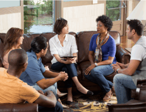 Six adults sit in a circle on couches and chairs, engaged in a group therapy session in a living room setting. One woman in the center holds a tablet and appears to be leading an effective healing discussion.