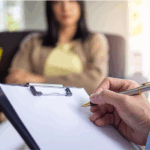 A psychiatrist holding a clipboard and pen takes notes while sitting across from a woman during a conversation, highlighting the role of psychiatrist in providing guidance during counseling sessions.