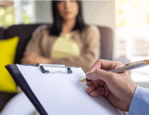 A psychiatrist holding a clipboard and pen takes notes while sitting across from a woman during a conversation, highlighting the role of psychiatrist in providing guidance during counseling sessions.