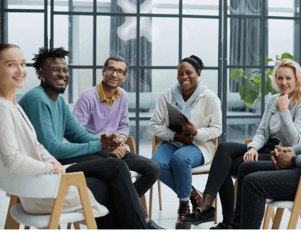 A diverse group of six adults sits together in a modern room, smiling and engaging in conversation as they share their experiences and Recovery journeys through IOP strategies.