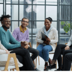 A diverse group of six adults sits together in a modern room, smiling and engaging in conversation as they share their experiences and Recovery journeys through IOP strategies.