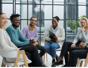 A diverse group of six adults sits together in a modern room, smiling and engaging in conversation as they share their experiences and Recovery journeys through IOP strategies.