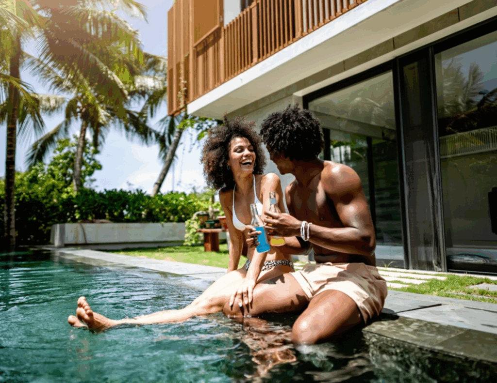 Two people sit by the edge of a pool outside a modern house, smiling and holding drinks, enjoying the tranquil setting—an ideal scene for couples retreats surrounded by palm trees and lush greenery.