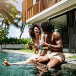 Two people sit by the edge of a pool outside a modern house, smiling and holding drinks, enjoying the tranquil setting—an ideal scene for couples retreats surrounded by palm trees and lush greenery.