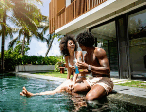 Two people sit by the edge of a pool outside a modern house, smiling and holding drinks, enjoying the tranquil setting—an ideal scene for couples retreats surrounded by palm trees and lush greenery.