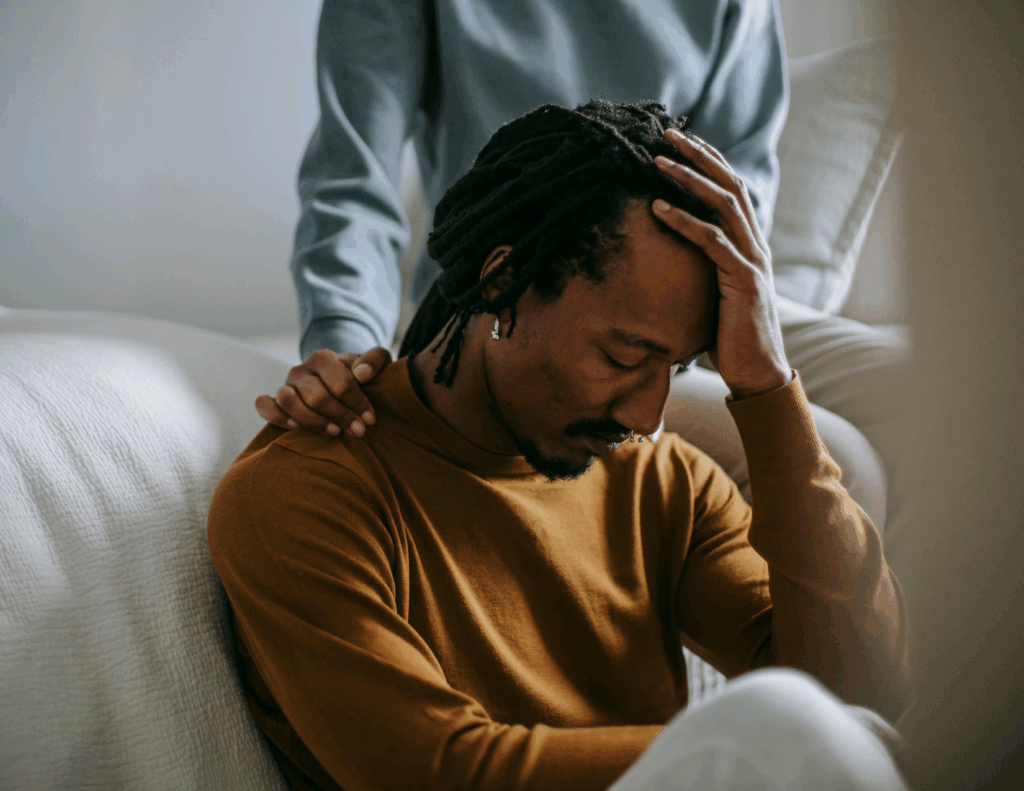 A person sits with their head in their hand, struggling with symptoms of complicated grief, while another offers support by resting a comforting hand on their shoulder.