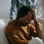 A person sits with their head in their hand, struggling with symptoms of complicated grief, while another offers support by resting a comforting hand on their shoulder.