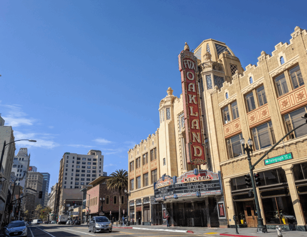 Art Deco-style theater with a vertical "Oakland" sign on a city street corner under a clear blue sky; nearby, signs point to a CBT Center offering trauma treatment. Surrounding buildings and street signs are visible.