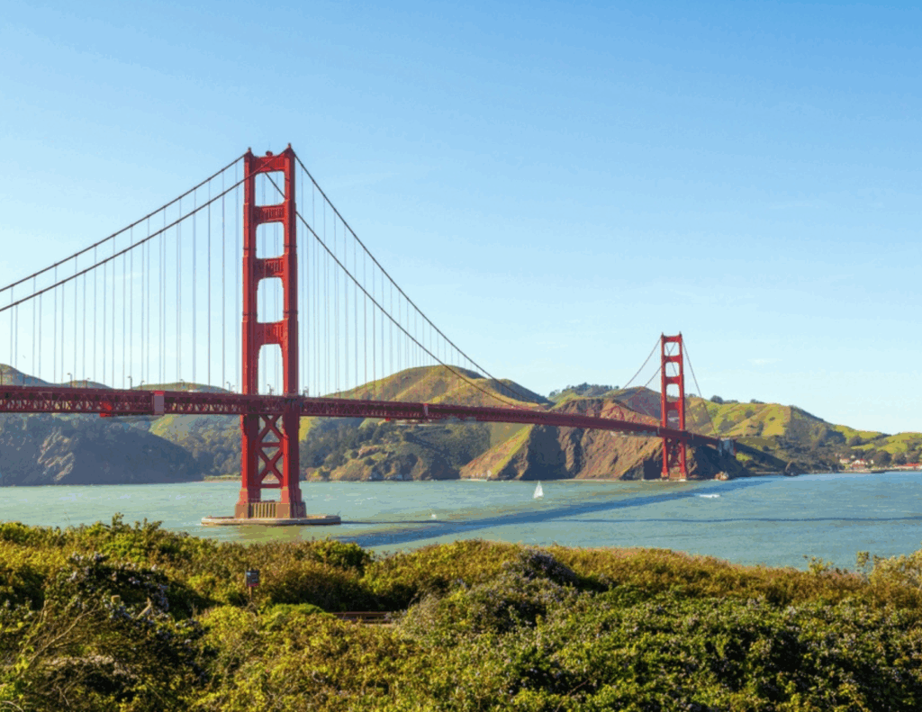 The Golden Gate Bridge spans across the blue water with rolling green hills in the background under a clear San Francisco sky.