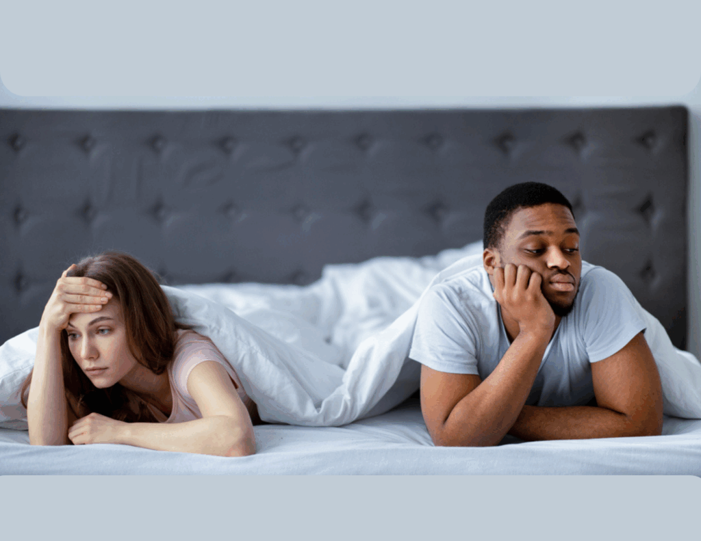 A woman and a man lie in bed under a white blanket, both looking upset and facing away from each other, suggesting relationship tension due to a mismatched sex drive.
