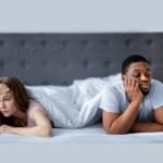 A woman and a man lie in bed under a white blanket, both looking upset and facing away from each other, suggesting relationship tension due to a mismatched sex drive.