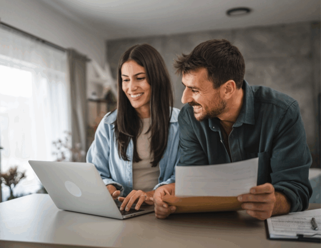 A woman using a laptop and a man holding papers sit together at a table, both smiling, in a bright home setting—discussing couples counseling and exploring their insurance coverage options.