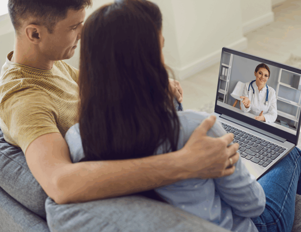A couple sits on a couch having a video call with a female doctor, using online couples therapy to transform their relationship, as she appears on the screen of their laptop.