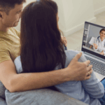 A couple sits on a couch having a video call with a female doctor, using online couples therapy to transform their relationship, as she appears on the screen of their laptop.