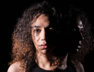A person with curly hair faces forward, half of their face brightly lit, while their profile and reflection—hinting at hidden lurking trauma—are cast in shadow against a dark background.