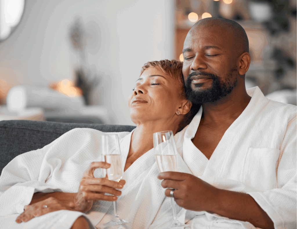 A couple wearing white robes relaxes on a couch with eyes closed, holding champagne glasses, in a cozy indoor setting—an ideal moment to reconnect and unwind at a couples wellness retreat.