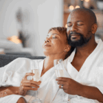 A couple wearing white robes relaxes on a couch with eyes closed, holding champagne glasses, in a cozy indoor setting—an ideal moment to reconnect and unwind at a couples wellness retreat.