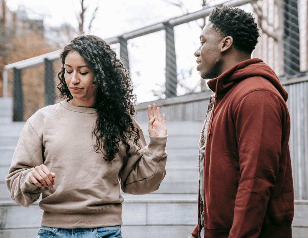 A woman with curly hair raises her hand dismissively—one of the clear signs marriage is over—while a man in a red hoodie stands nearby on outdoor steps.