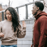 A woman with curly hair raises her hand dismissively—one of the clear signs marriage is over—while a man in a red hoodie stands nearby on outdoor steps.