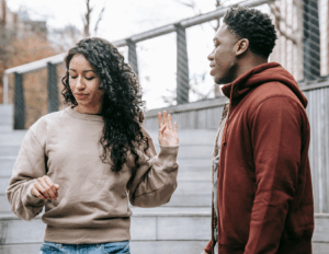 A woman with curly hair raises her hand dismissively—one of the clear signs marriage is over—while a man in a red hoodie stands nearby on outdoor steps.