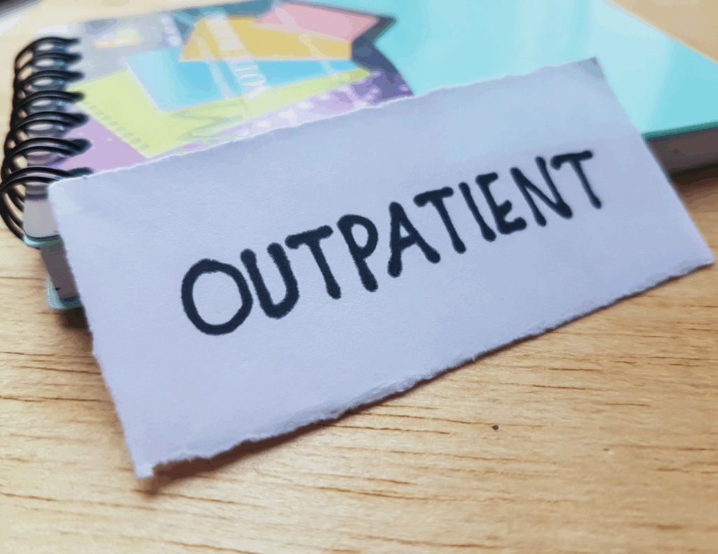 A piece of paper with the word "OUTPATIENT" handwritten in black marker rests on a wooden surface near a spiral notebook, suggesting a connection to comprehensive treatment solutions or IOP mental health programs.