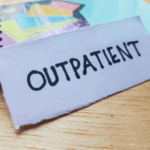 A piece of paper with the word "OUTPATIENT" handwritten in black marker rests on a wooden surface near a spiral notebook, suggesting a connection to comprehensive treatment solutions or IOP mental health programs.