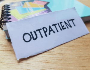 A piece of paper with the word "OUTPATIENT" handwritten in black marker rests on a wooden surface near a spiral notebook, suggesting a connection to comprehensive treatment solutions or IOP mental health programs.