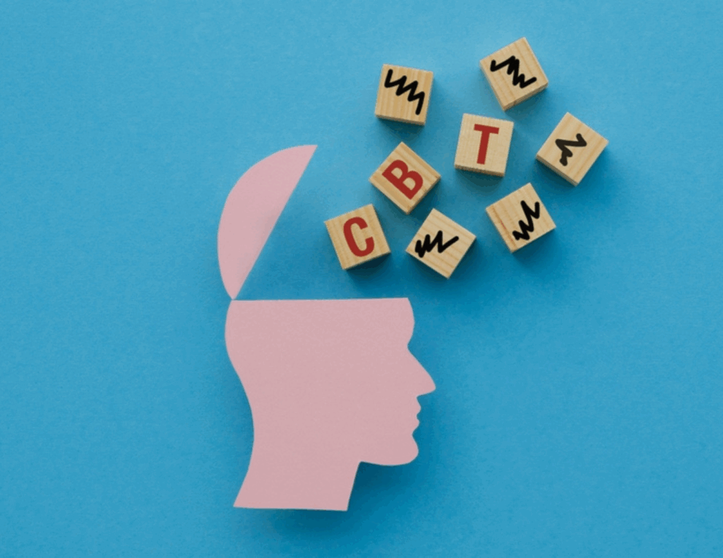 A pink paper cutout of a human head with an open top sits next to wooden blocks spelling "CBT," symbolizing mental health and mindfulness on a blue background.