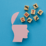 A pink paper cutout of a human head with an open top sits next to wooden blocks spelling "CBT," symbolizing mental health and mindfulness on a blue background.