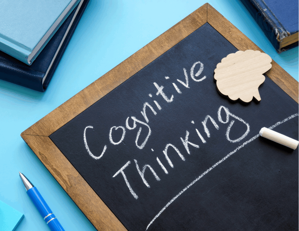 A chalkboard with "Cognitive Thinking" written on it is surrounded by books, a wooden brain-shaped cutout, a piece of chalk, and a blue pen—depicting the tools used to understand thinking and identify cognitive distortions.