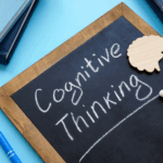 A chalkboard with "Cognitive Thinking" written on it is surrounded by books, a wooden brain-shaped cutout, a piece of chalk, and a blue pen—depicting the tools used to understand thinking and identify cognitive distortions.