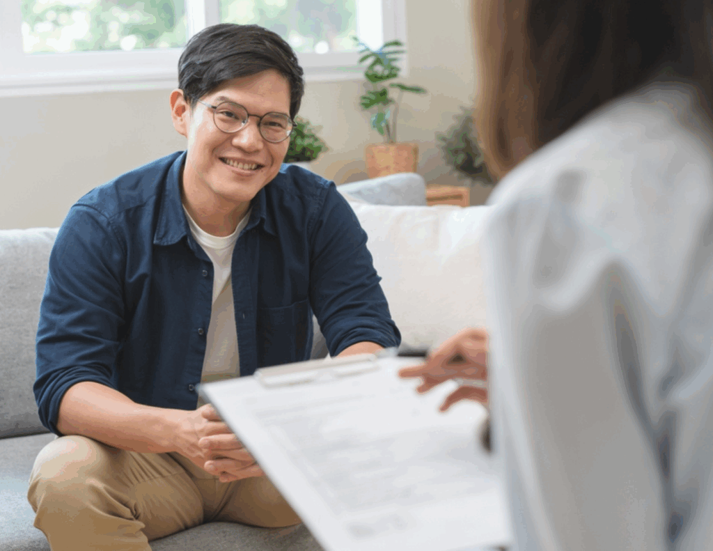 A man sits on a couch smiling while talking to a psychiatrist holding a clipboard during a consultation in a bright room, highlighting the importance of seeking help for mental health.