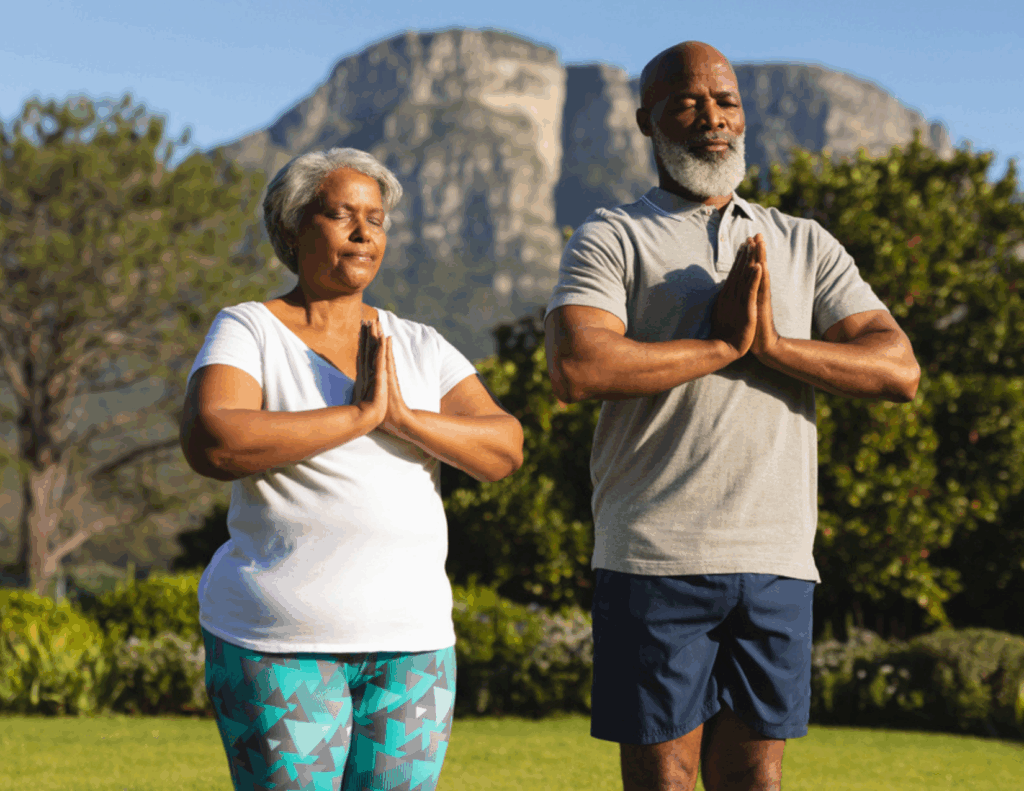 Two older adults stand outdoors with hands together in a prayer pose, practicing yoga in front of a mountain and greenery—an uplifting moment often found at marriage retreats.