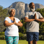 Two older adults stand outdoors with hands together in a prayer pose, practicing yoga in front of a mountain and greenery—an uplifting moment often found at marriage retreats.
