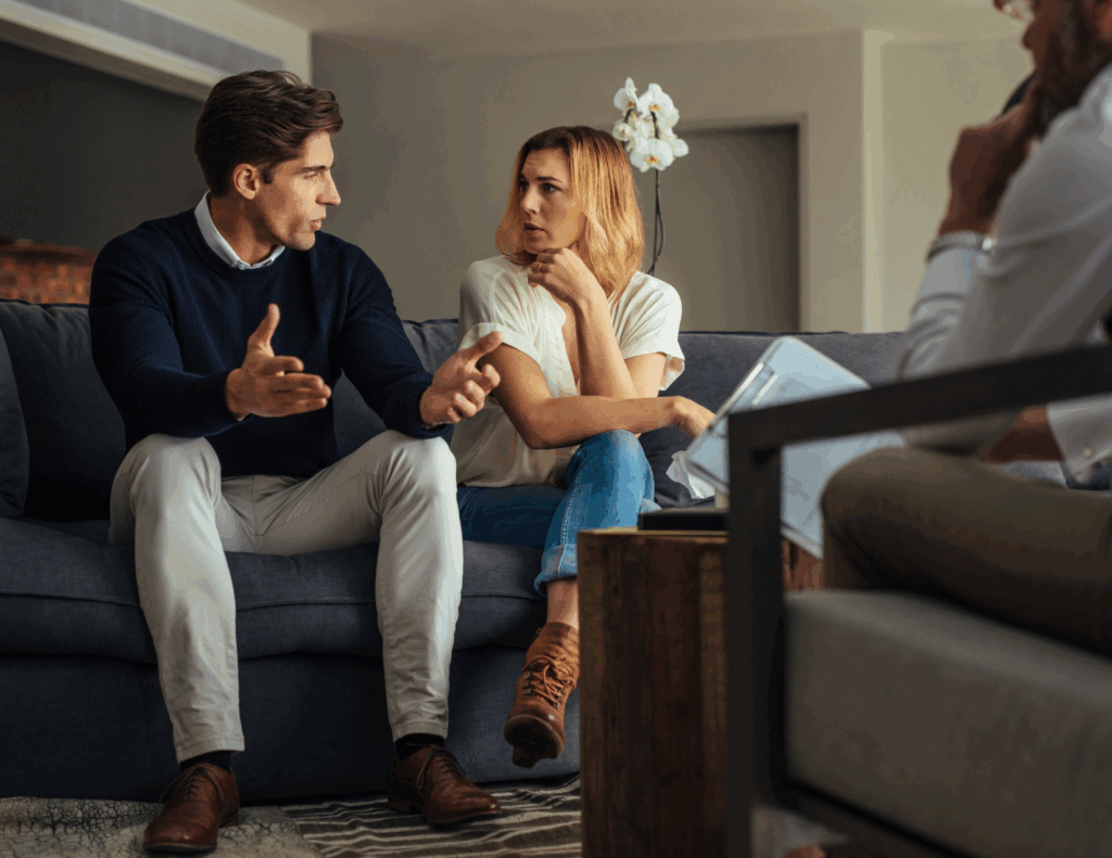 A man and woman sit on a couch talking, with a third person nearby holding a clipboard, suggesting a couples counseling session.