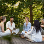 Five adults sit together on a wooden deck outdoors, engaged in conversation at a mental health retreat, surrounded by greenery, with tea and snacks on the ground.
